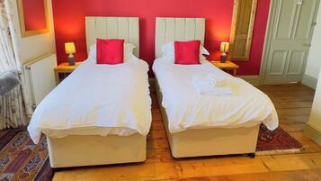 A room with two single beds, each with a red pillow and white bedding. There are two bedside tables with lamps, a radiator, and a patterned rug on the floor.