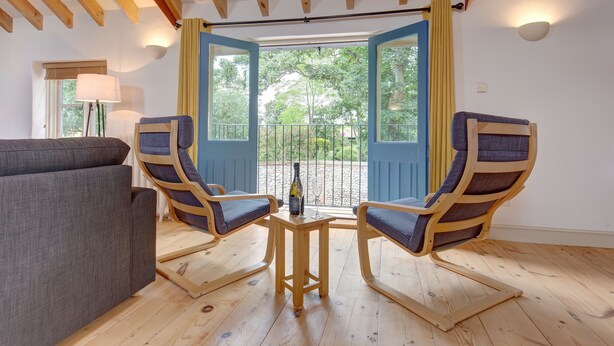 A room with a wooden ceiling, a grey sofa, two wooden chairs with cushions, a small wooden table with a bottle and glass, and a view through an open door to the outside. - Owl Barn -  a self catering cottage that sleeps 6 guests  in 3 bedrooms (Morston)