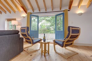 Ein Zimmer mit Holzboden, einem grauen Sofa, zwei Holzsesseln mit Kissen, einem kleinen Holztisch mit Flasche und Glas sowie Blick durch eine offene Tür ins Freie.
