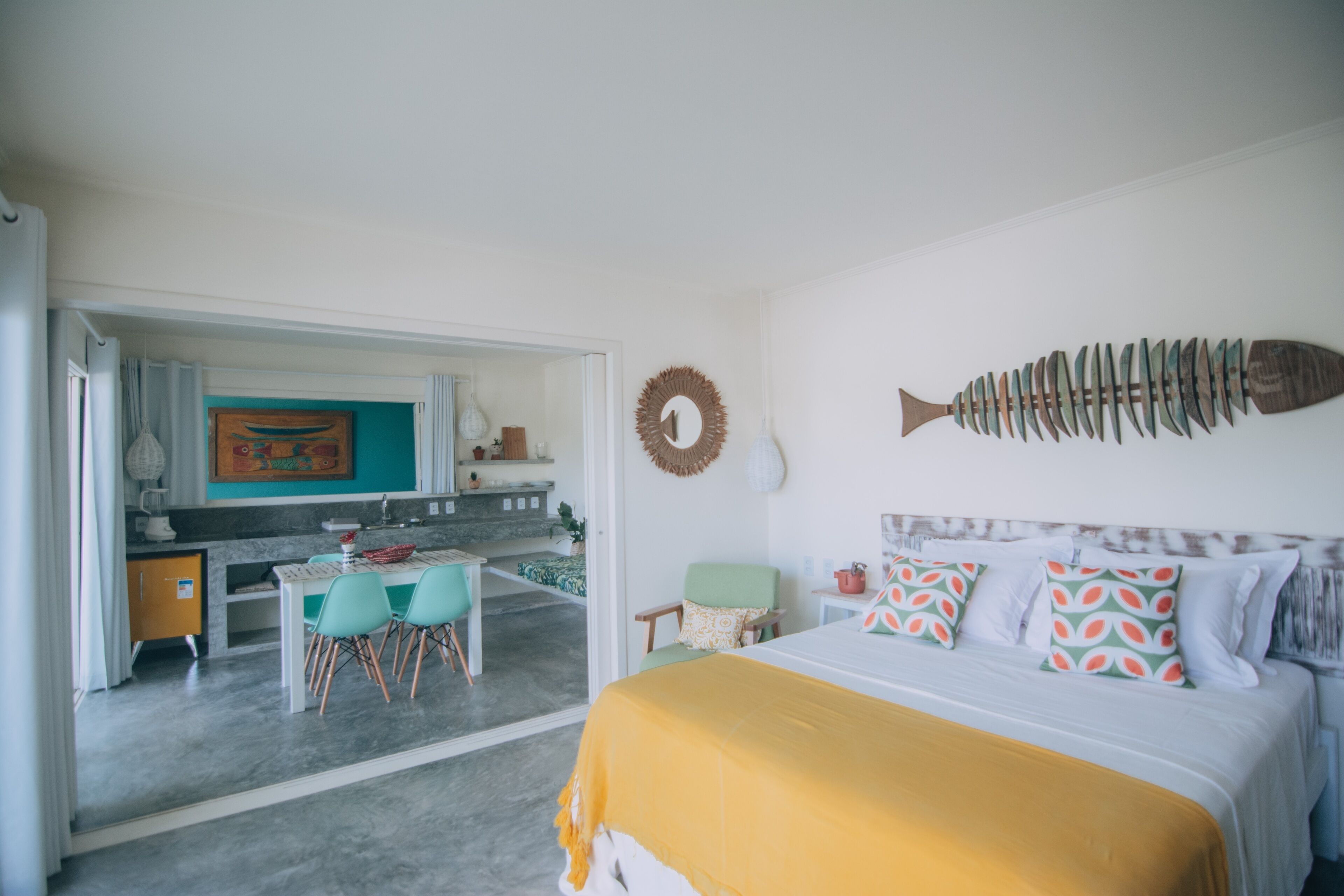 Ein Schlafzimmer mit Bett, einer Fisch-Wanddekoration, einem Essbereich mit Stühlen und einer durchgehenden Tür erkennbaren Küche.