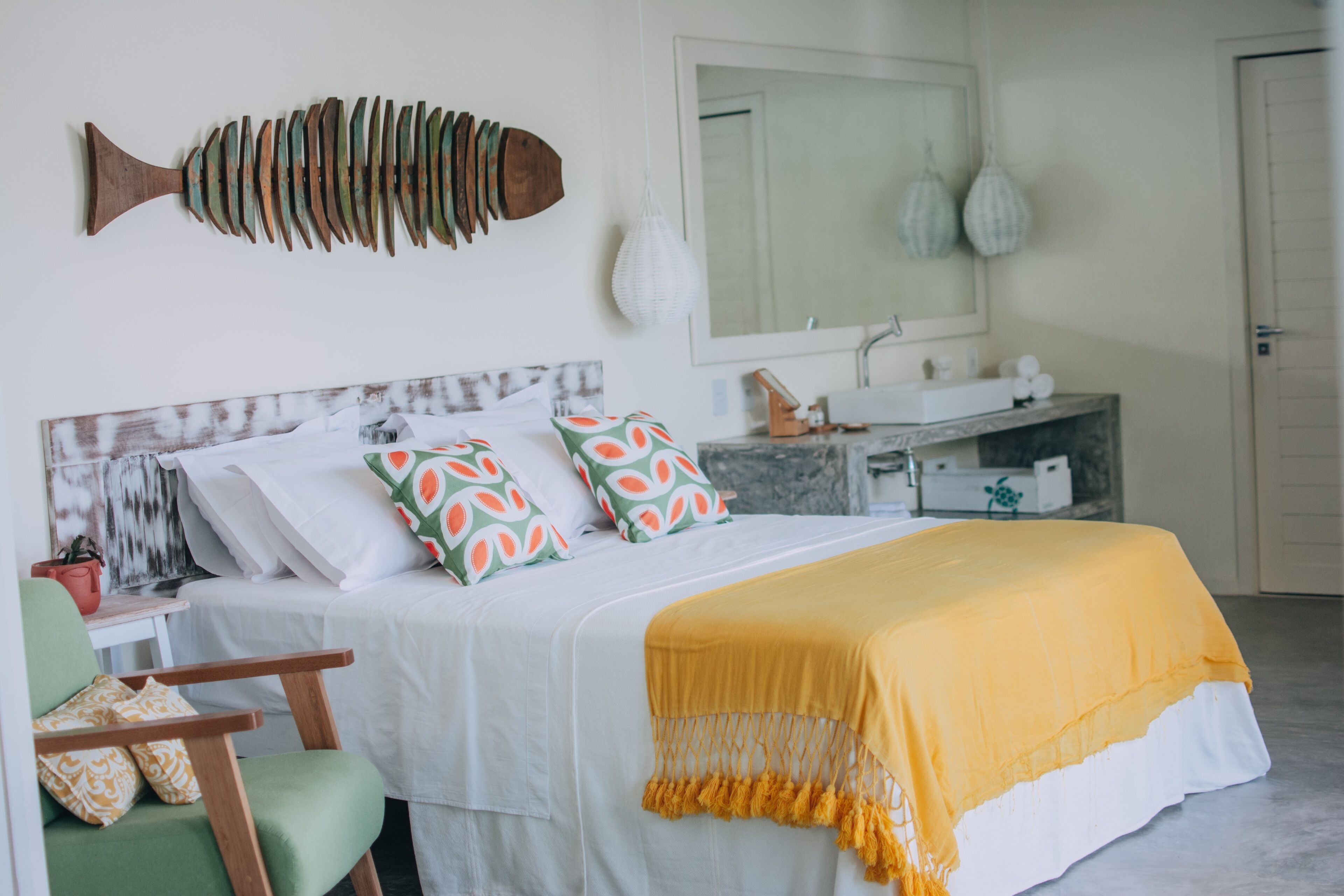 A bedroom with a bed, a green chair, a wooden fish wall art, a mirror, and a sink.