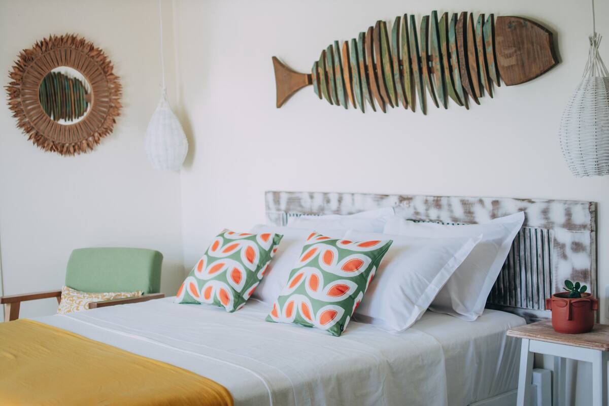 Un dormitorio ordenado con cama, almohadas, una silla verde, una mesita con una planta y adornos de pared, incluyendo una pieza con forma de pez y un espejo circular.