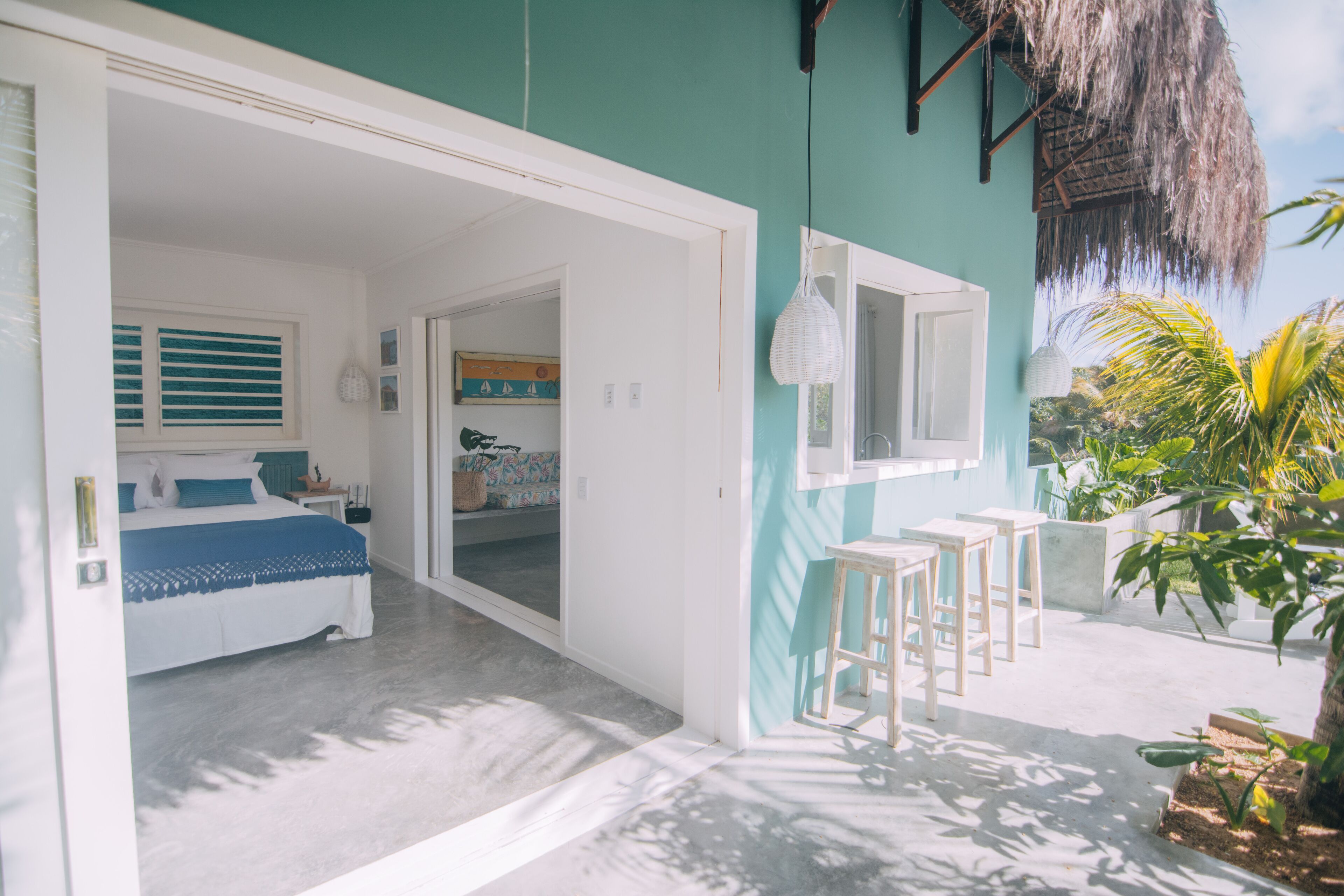 Une chambre à coucher avec un lit, une fenêtre avec des stores, une porte donnant sur une autre pièce et un balcon doté d’un toit de chaume et de plantes en pot.