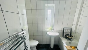 Baño pequeño con lavamanos, inodoro y bañera blancos. Una ventana ofrece luz natural y se ve un letrero enmarcado en la pared.