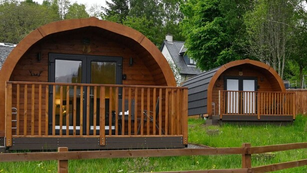 Deux cabanes en bois, dotées de toits en arc et de balustrades en bois, sont entourées d’une zone herbeuse parsemée de fleurs sauvages.