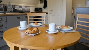 Une table à manger avec des assiettes de croissants et des tasses de café, une chaise en bois et, au fond, une cuisine.