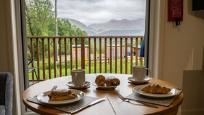 Une table ronde en bois, sur laquelle sont disposés des aliments pour le petit-déjeuner, notamment des croissants et une assiette de nourriture, le tout placé devant un paysage de montagnes et de verdure, visible à travers la rambarde d’un balcon.