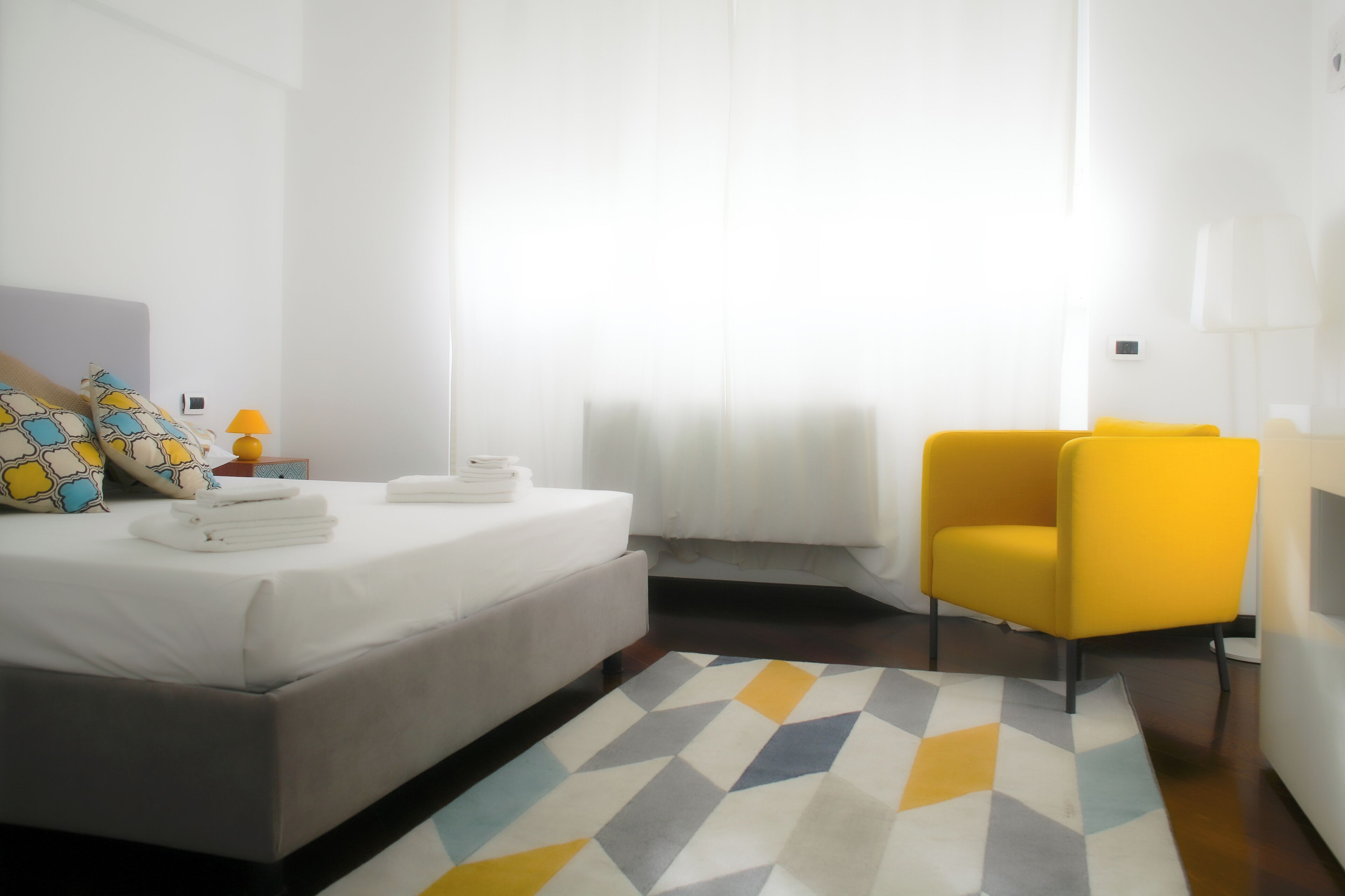 Una habitación de hotel moderna con una cama, una mesita de noche con lámpara, un sillón amarillo y una alfombra de estampado geométrico.