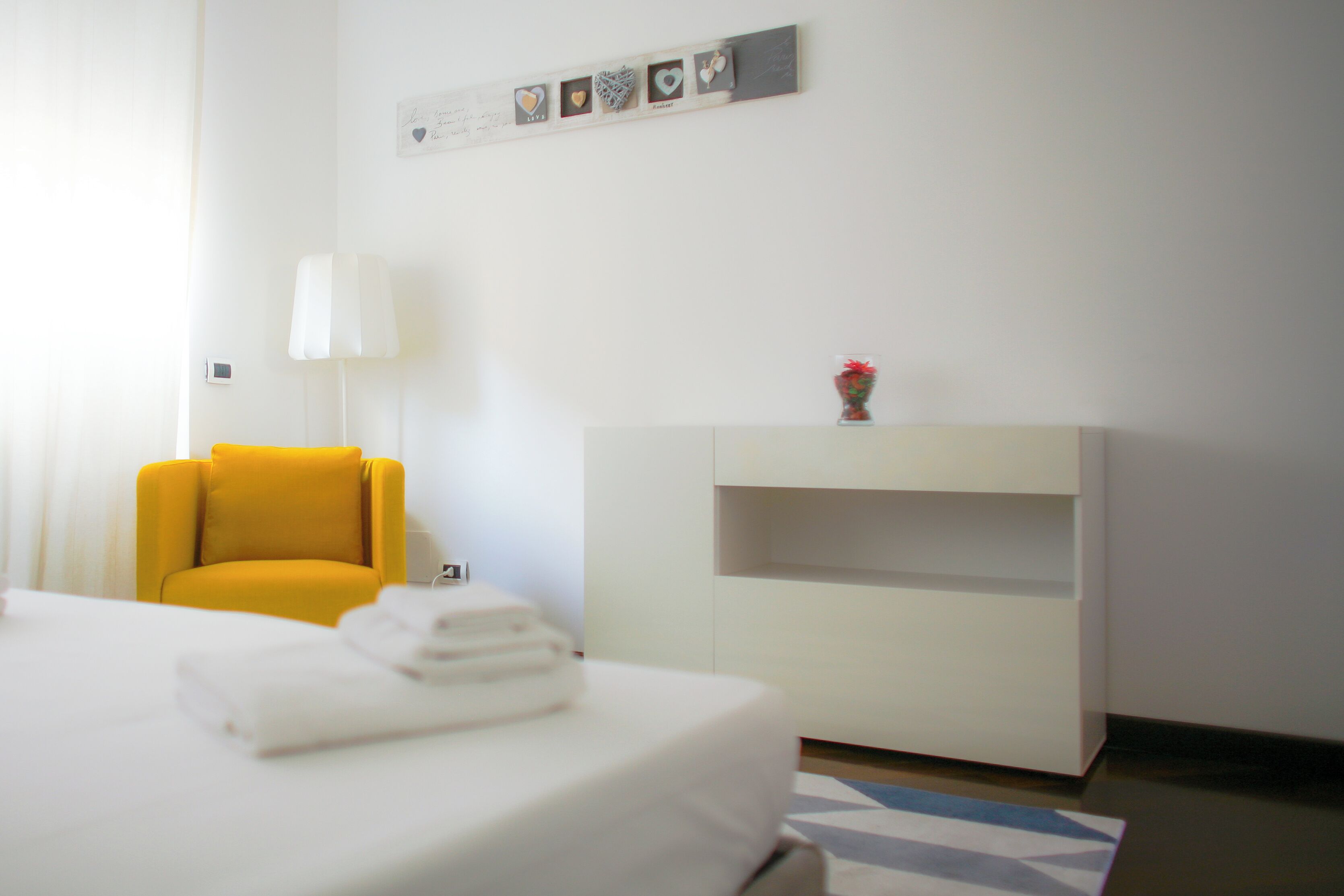 A hotel room with a bed, a yellow armchair, a white bedside table, a lamp, and a wall-mounted shelf with a vase.