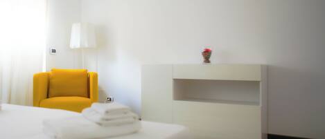 A hotel room with a bed, a yellow armchair, a white bedside table, a lamp, and a wall-mounted shelf with a vase.