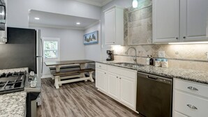 A modern kitchen with white cabinetry, a granite countertop, a built-in dishwasher, and a window with blinds.