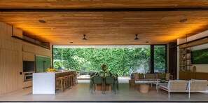 A modern kitchen with a central island, bar stools, and a dining area with a view of lush greenery.