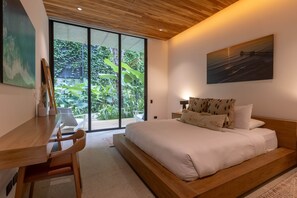 A modern bedroom with a large bed, a desk, and a view of greenery through a sliding door.