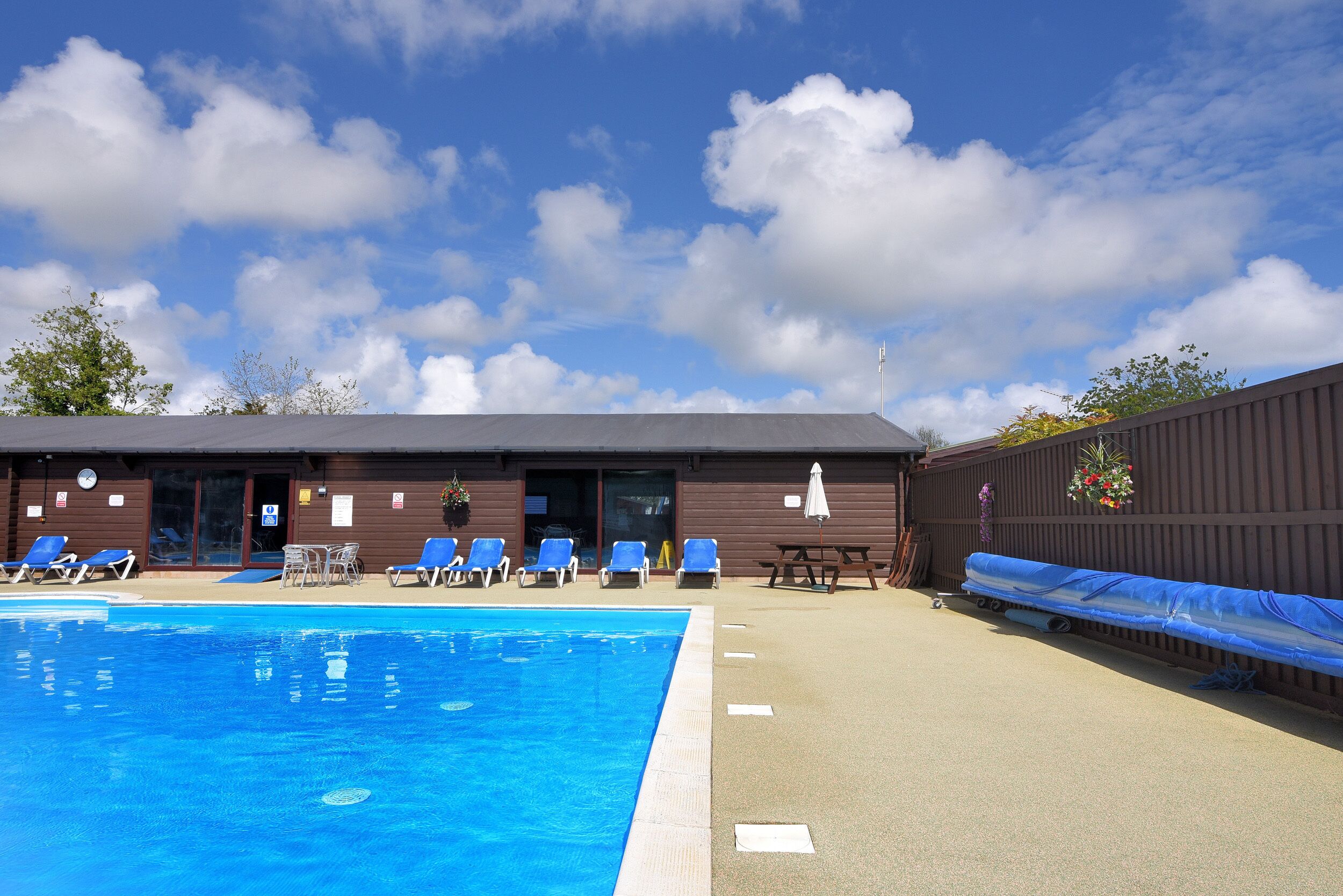 Uma piscina com cadeiras azuis e uma área coberta, um edifício com fachada marrom, e um céu claro com nuvens.