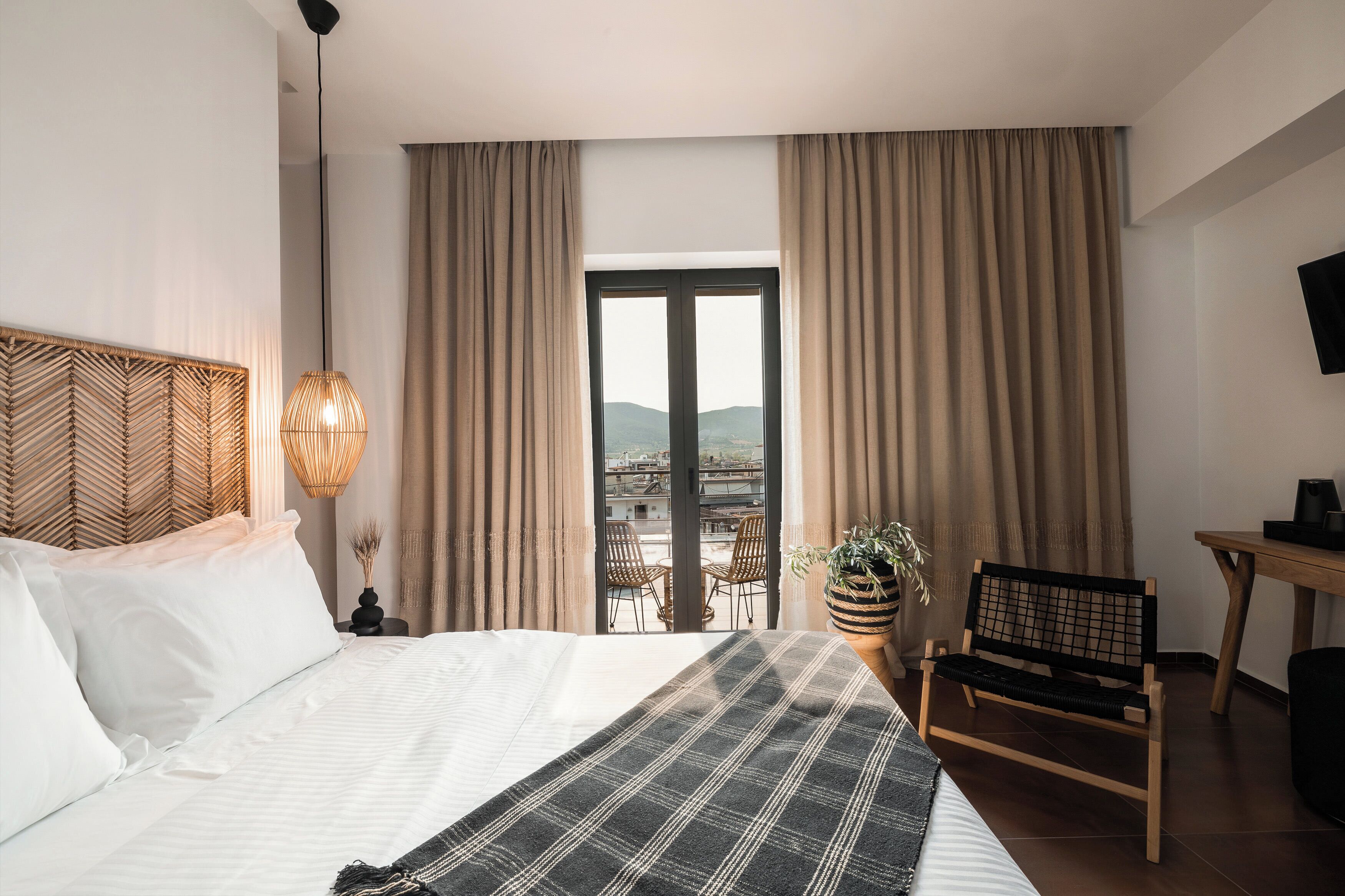 A hotel room with a bed, a desk, a chair, a potted plant, and a view of the city through the window.
