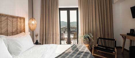 A hotel room with a bed, a desk, a chair, a potted plant, and a view of the city through the window.