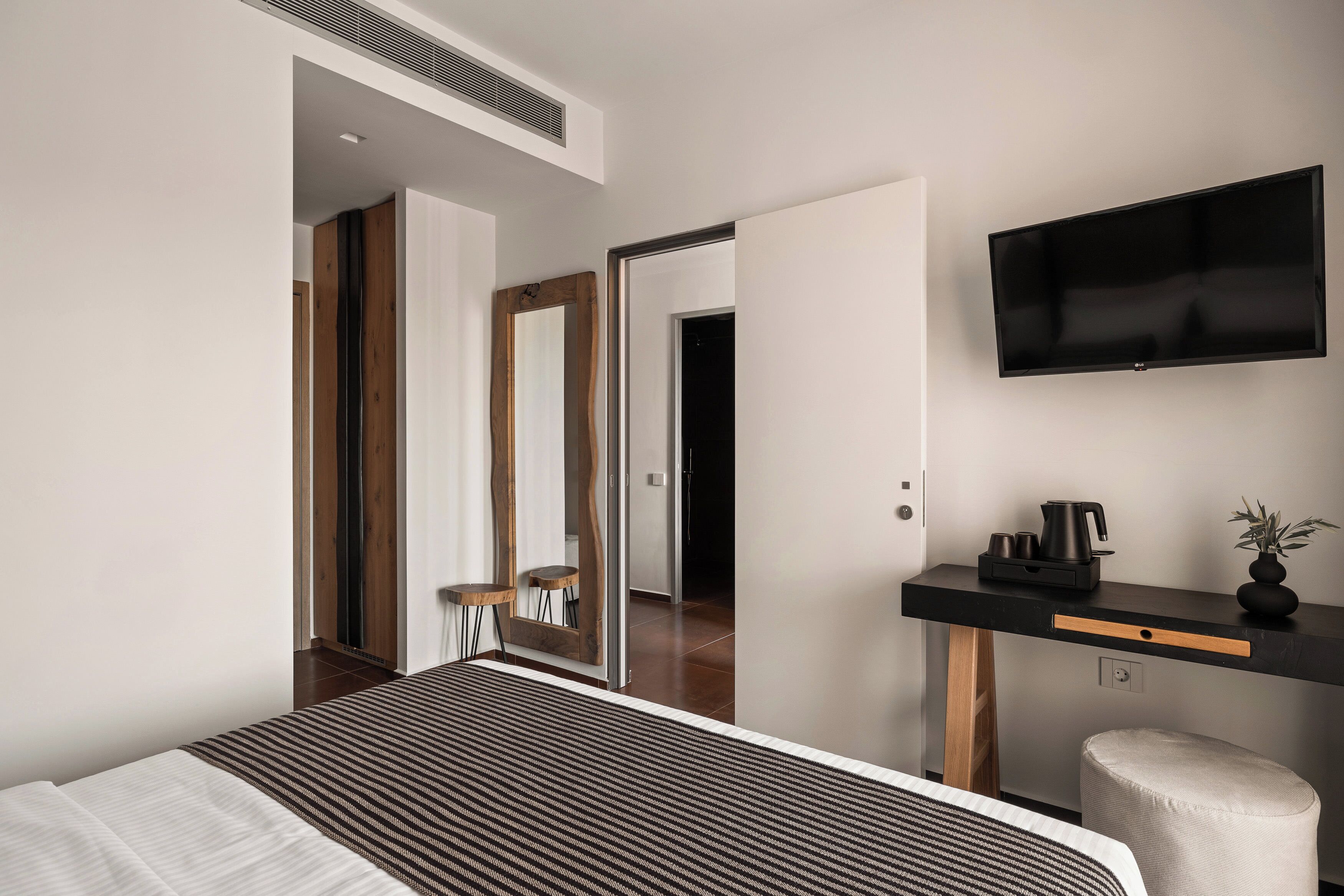 A modern hotel room with a large bed, a flat-screen TV mounted on the wall, a small desk with a coffee maker, and a mirror.
