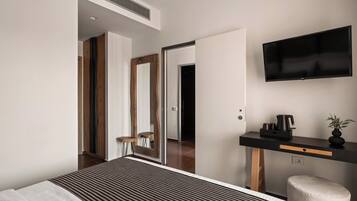 A modern hotel room with a large bed, a flat-screen TV mounted on the wall, a small desk with a coffee maker, and a mirror.