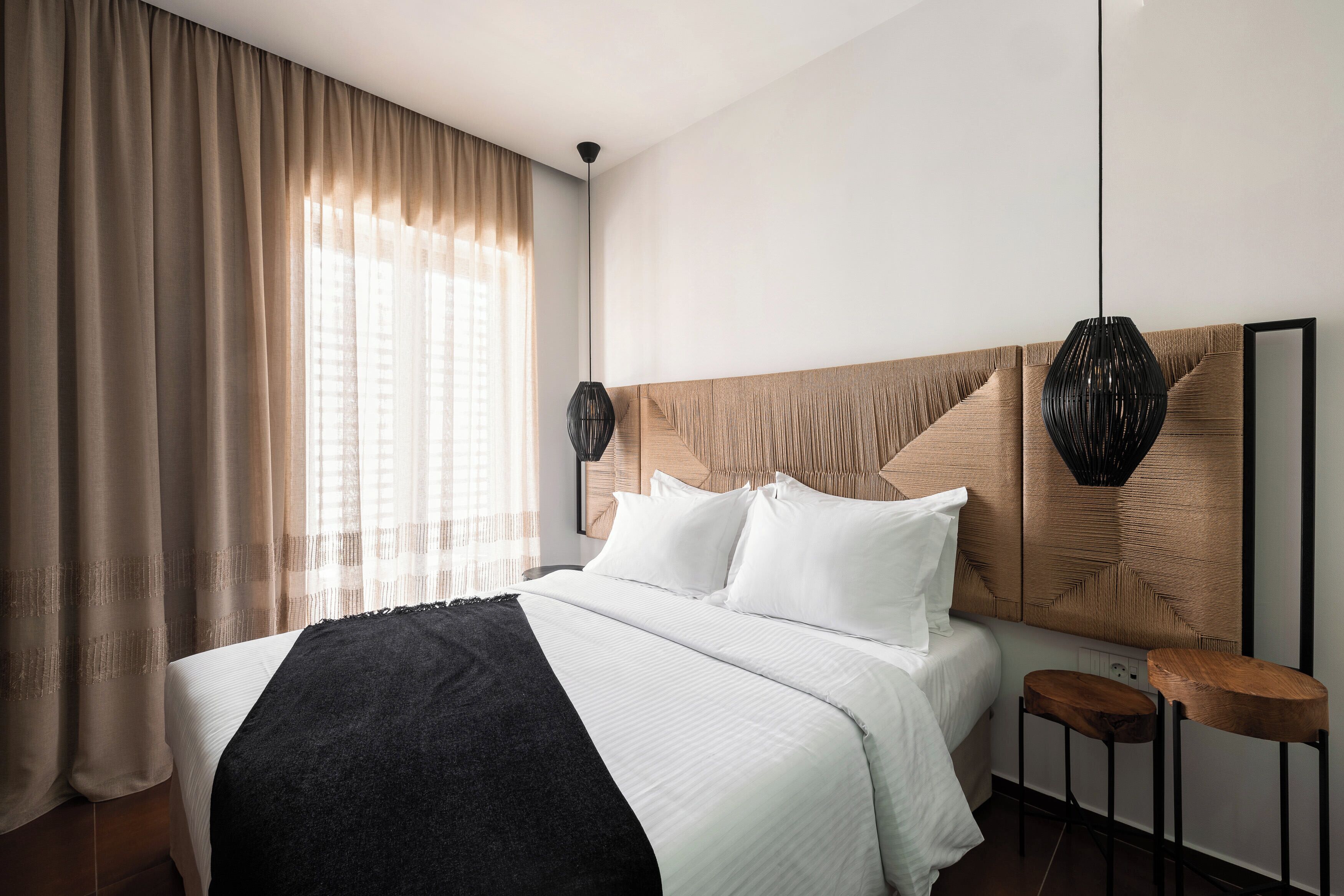 Une chambre d’hôtel avec un grand lit, une tête de lit en bois, deux lampes suspendues et un petit tabouret en bois.