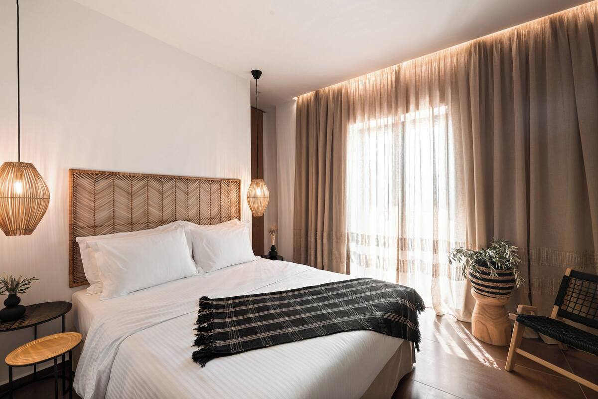 Una habitación de hotel moderna con una cama grande, una mesita de noche, una silla, una planta en maceta y un ventanal con cortinas.