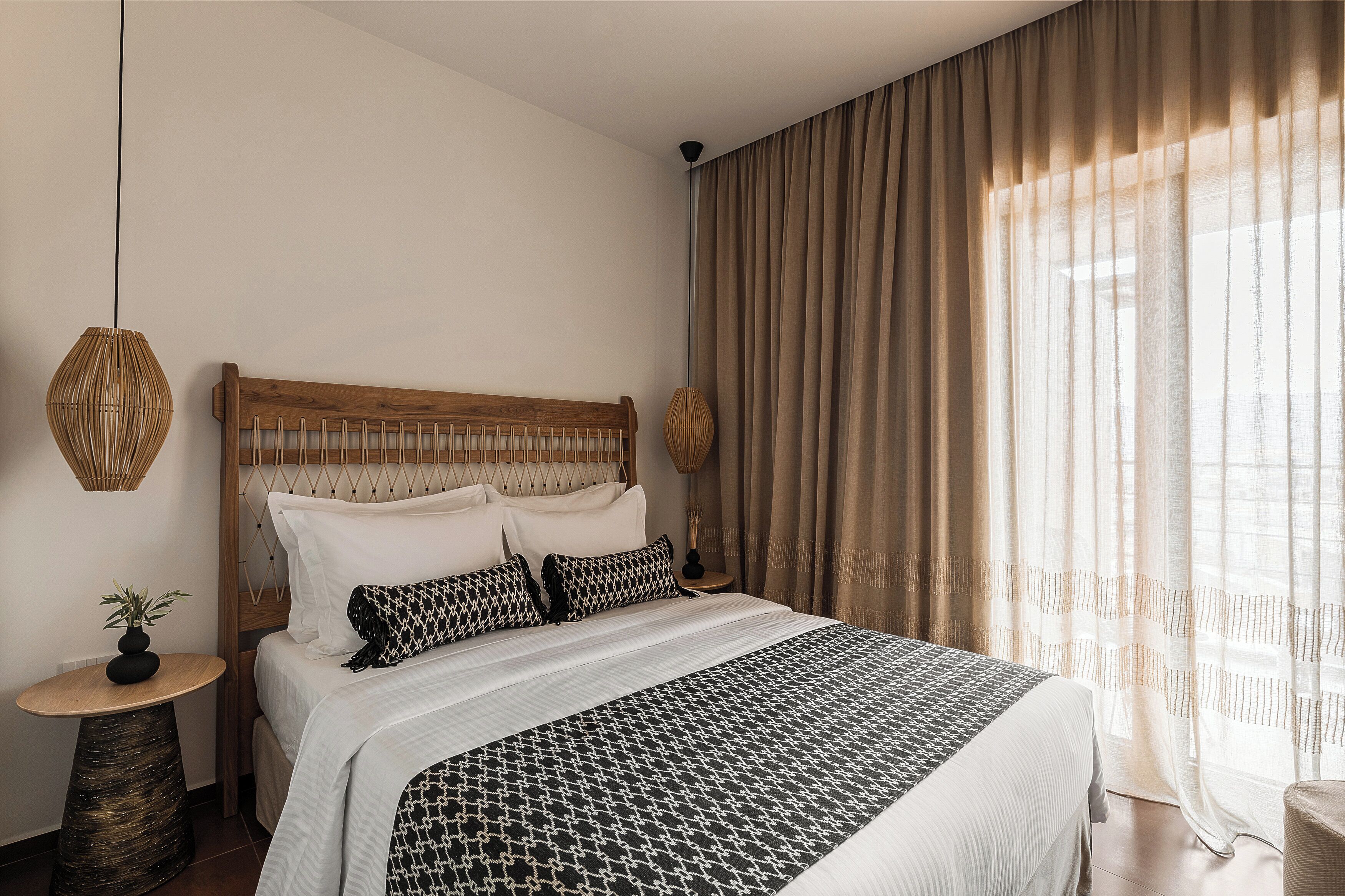 A neatly arranged bedroom with a wooden headboard, a large bed with patterned bedding, a bedside table with a lamp, and sheer curtains allowing natural light.