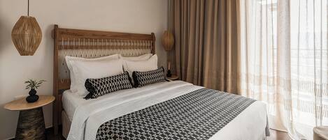 Une chambre à coucher bien aménagée, avec une tête de lit en bois, un grand lit recouvert d’une literie à motifs, une table de chevet avec une lampe et des rideaux légers qui laissent passer la lumière naturelle.