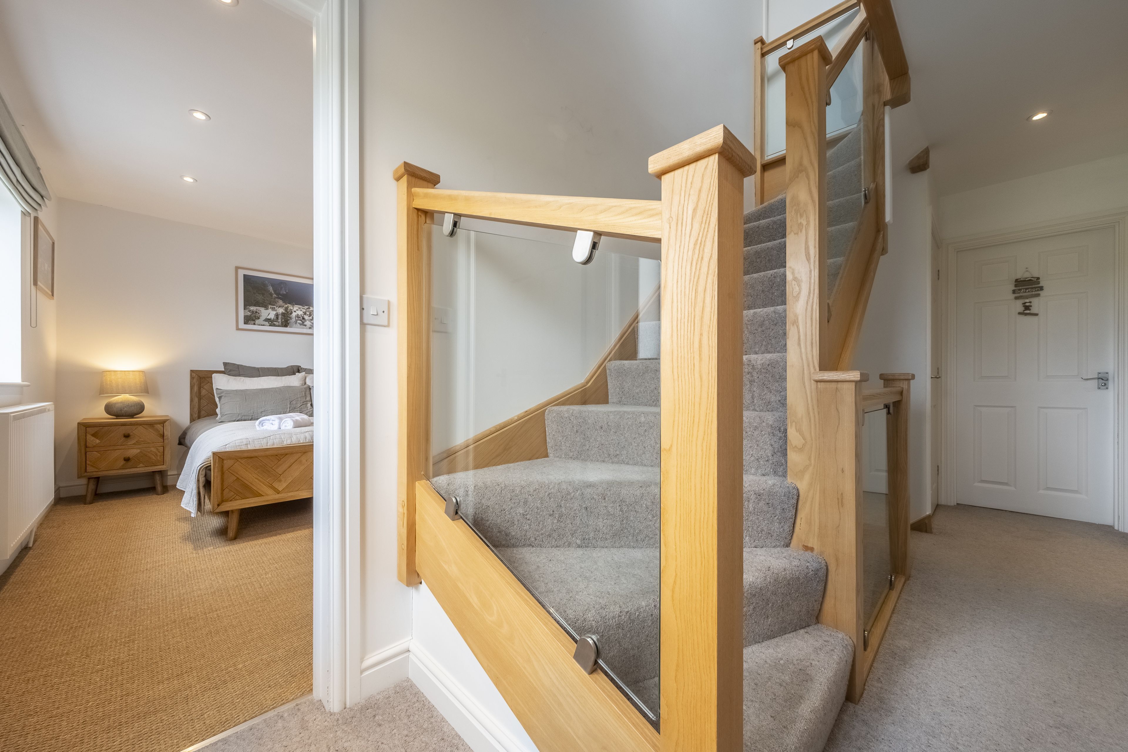 Eine Treppe mit Holzgeländern und Teppichstufen führt zu einem Schlafzimmer mit Bett, Nachttisch und einem an der Wand hängenden Bild.