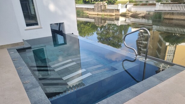 Una piscina infinita moderna con borde de cristal, rodeada de baldosas color beige y un borde de piedra.
