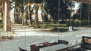 Un espacio exterior para comer con mesas y sillas, ventanas amplias y vistas a árboles y un sendero.