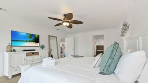 Ein Schlafzimmer mit einem großen Bett, einem Deckenventilator, einem Fernseher, der eine Strandansicht zeigt, und einer weißen Kommode.