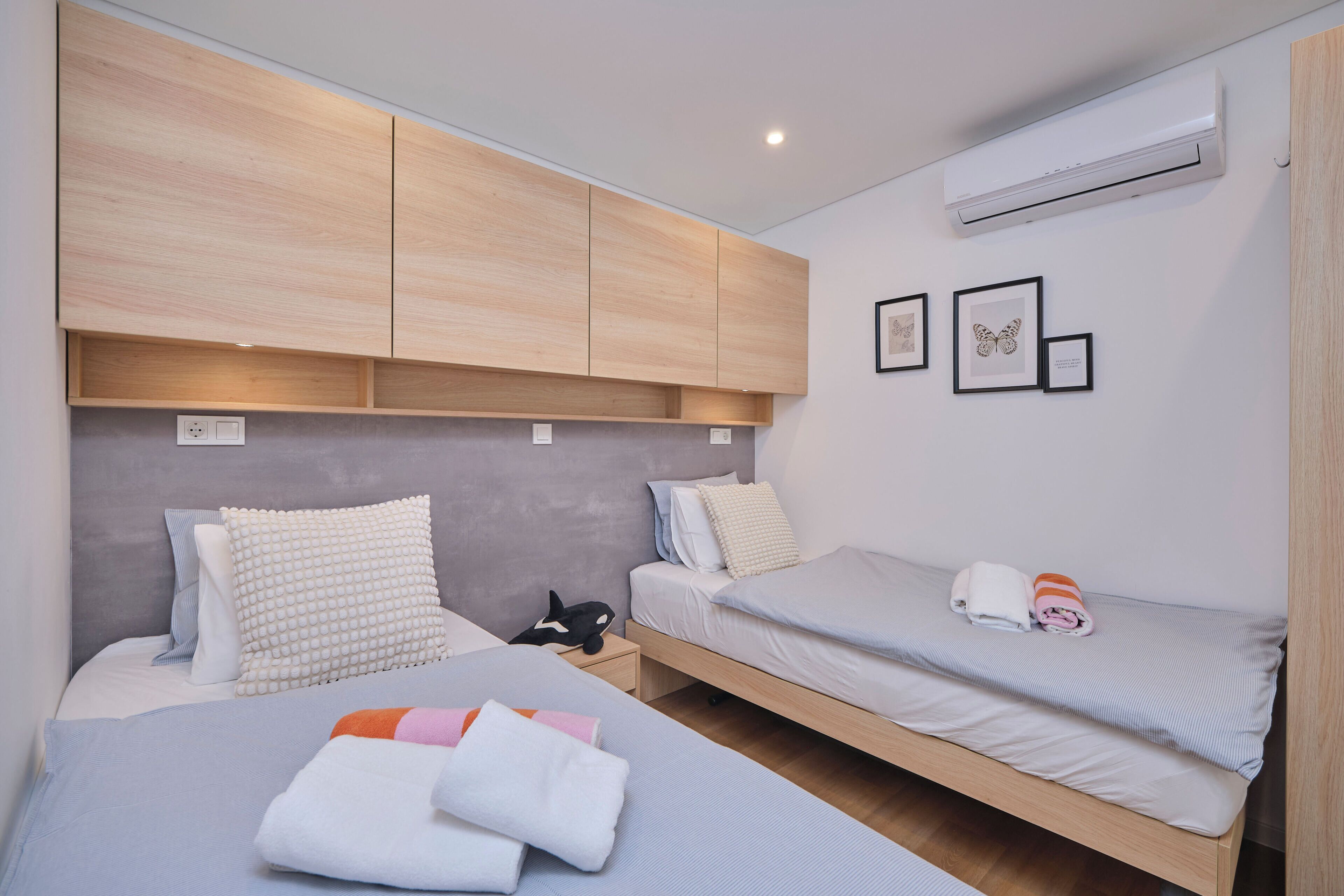 A bedroom with two beds, a wardrobe, an air conditioner, and framed pictures on the wall.