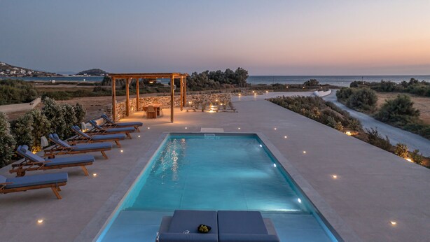 A poolside area with lounge chairs, a pergola, and a view of the sea at dusk.