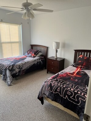 A bedroom with two beds, each with a Star Wars themed bedspread, a ceiling fan, a bedside table with a lamp, and a window with blinds.
