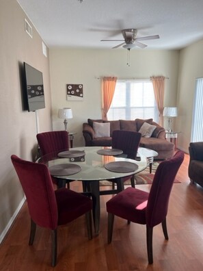 A living room with a round glass table, red chairs, a brown sofa, and a ceiling fan.
