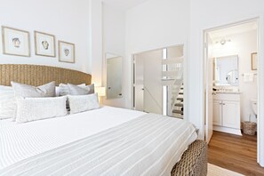 A bedroom with a large bed, a wicker headboard, and a view of a staircase leading to a bathroom. - Oceanfront, 4 BR/5 BA, EV Charging, Super Stylish (Hilton Head Island)