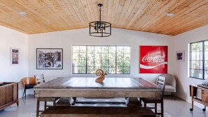 A dining area with a wooden table, chairs, a bench, a ceiling light fixture, and a large window with a view of trees.