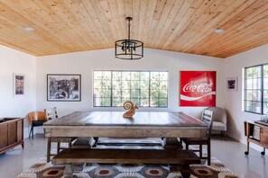 A dining area with a wooden table, chairs, a bench, a ceiling light fixture, and a large window with a view of trees.