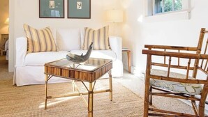 A living room with a white sofa, striped pillows, a glass coffee table with a bird sculpture, a wooden chair, and two framed pictures on the wall. - Historic 2BR Nantucket Cottage | Walk to Downtown (Nantucket)