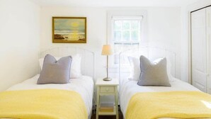 A room with two beds, each with a white headboard and a yellow blanket. There is a nightstand with a lamp between the beds. A painting of a landscape hangs on the wall above the beds. - Historic 2BR Nantucket Cottage | Walk to Downtown (Nantucket)