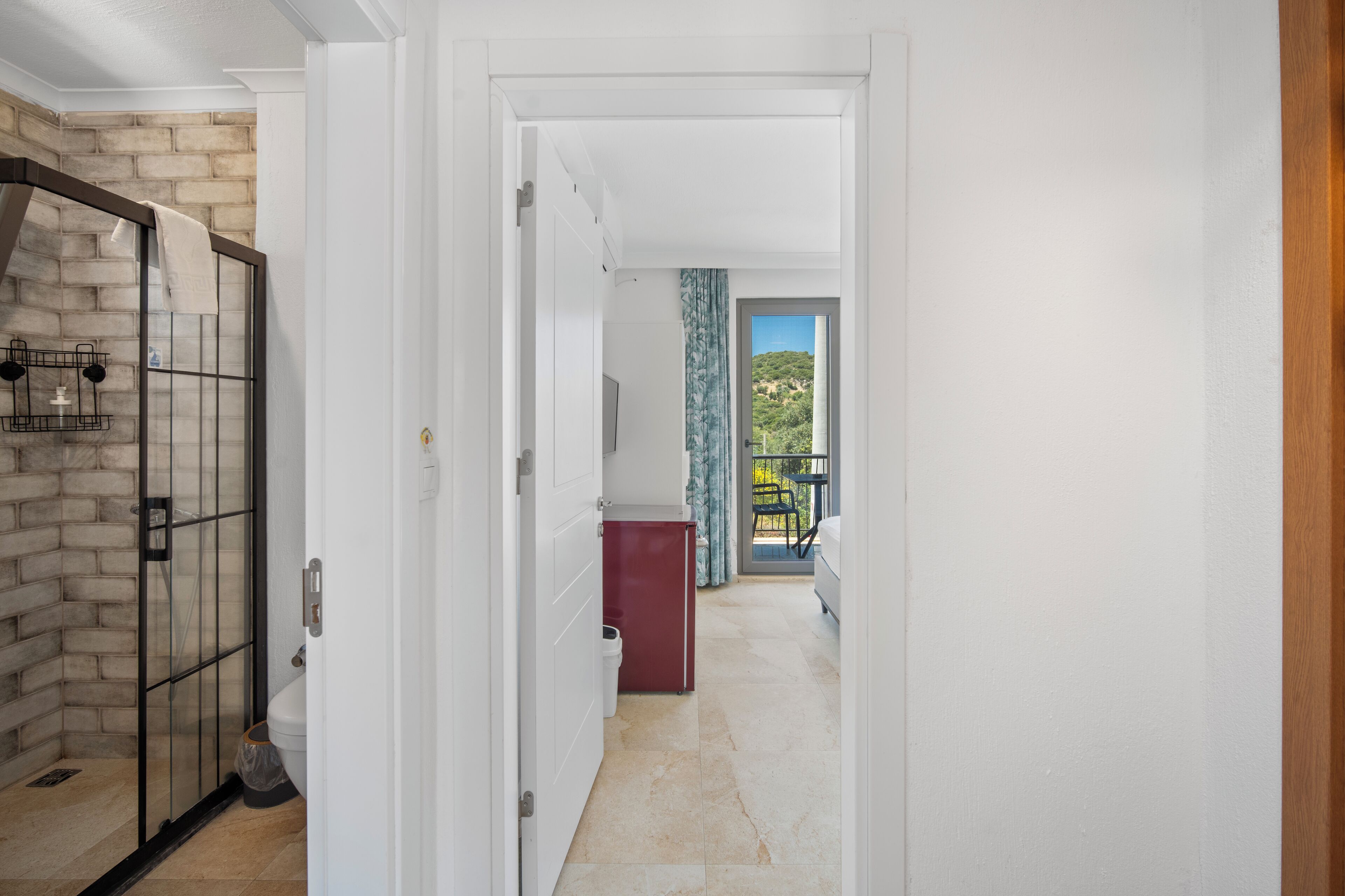 A hallway with a glass door leading to a bathroom, a red cabinet, and a view of a balcony with a chair.