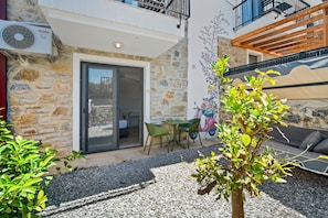 A stone building with a sliding glass door, a small patio with a table and chairs, and a garden area with a tree.