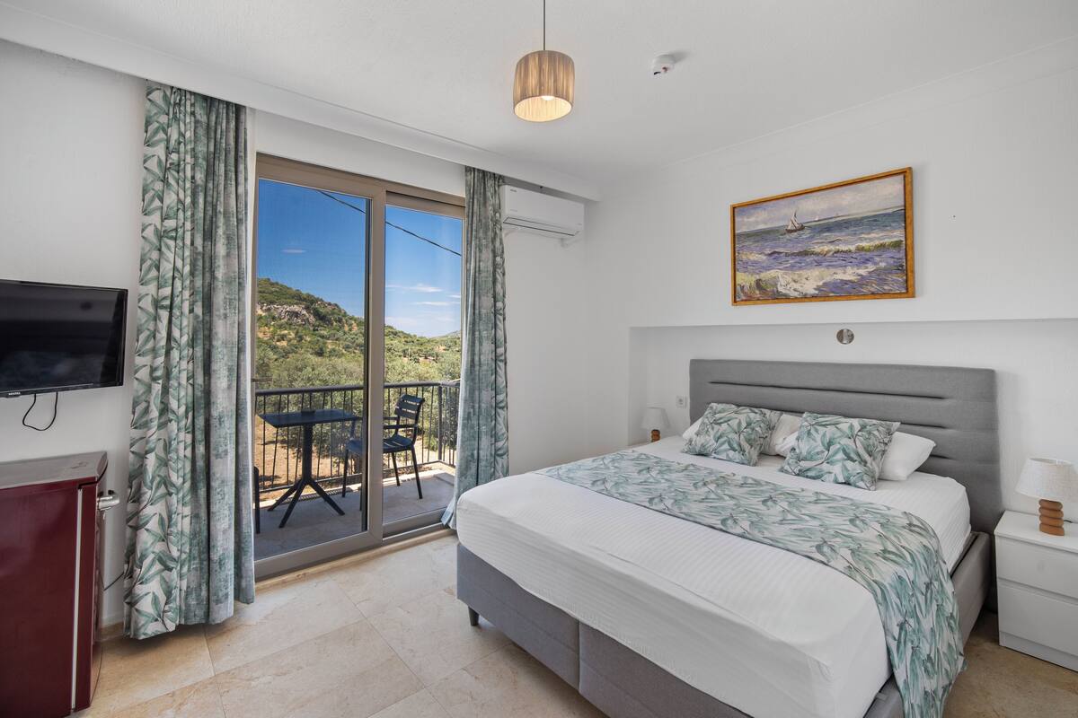 A bedroom with a bed, a TV, a painting, and a balcony with a view of the sea.