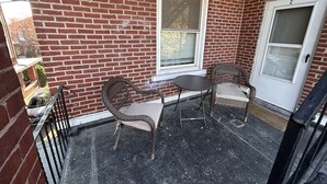 A covered porch with a brick wall, two wicker chairs, a small round table, and a door marked with the number 2.