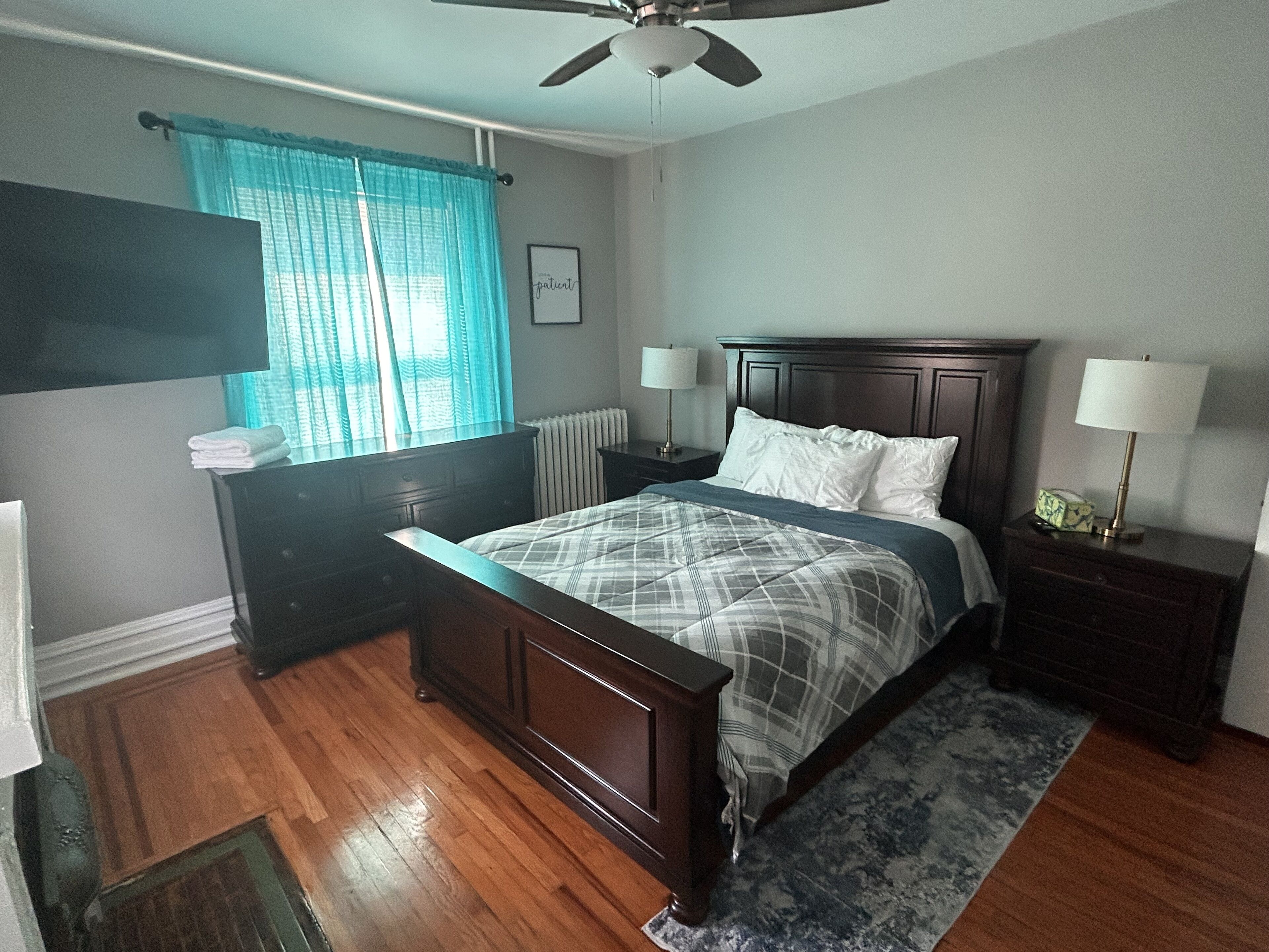 A bedroom with a bed, nightstands, a dresser, a ceiling fan, and a window with curtains.