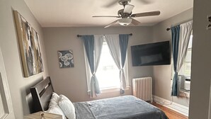 A bedroom with a bed, a ceiling fan, a TV mounted on the wall, and a window with curtains.
