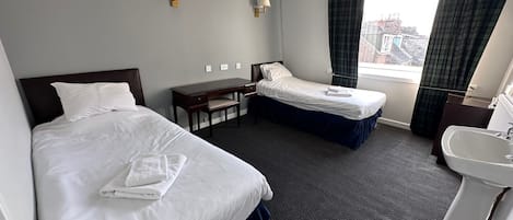 A hotel room with two beds, a desk, a chair, a small table, a sink, and a window with curtains.