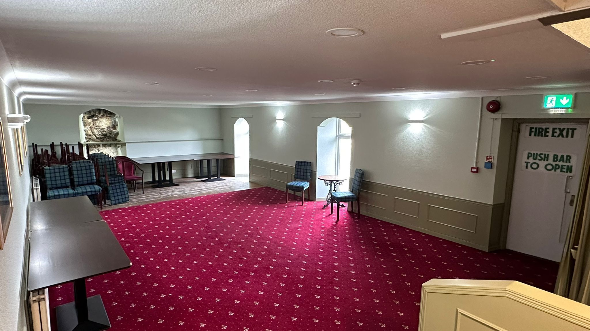Une pièce spacieuse avec une moquette rouge, plusieurs chaises, une table et une porte de sortie incendie équipée d’une barre de poussée.