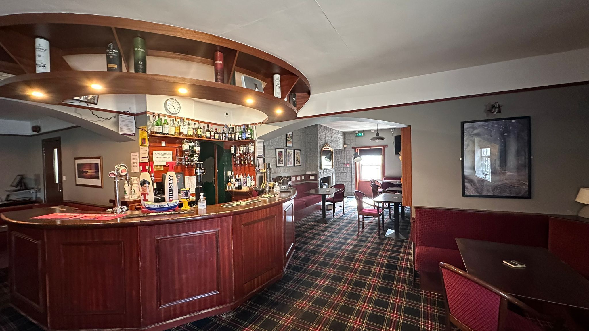 A bar area with a curved wooden counter, a variety of liquor bottles, and a clock on the wall. There are seating areas with plaid flooring and red upholstered chairs.