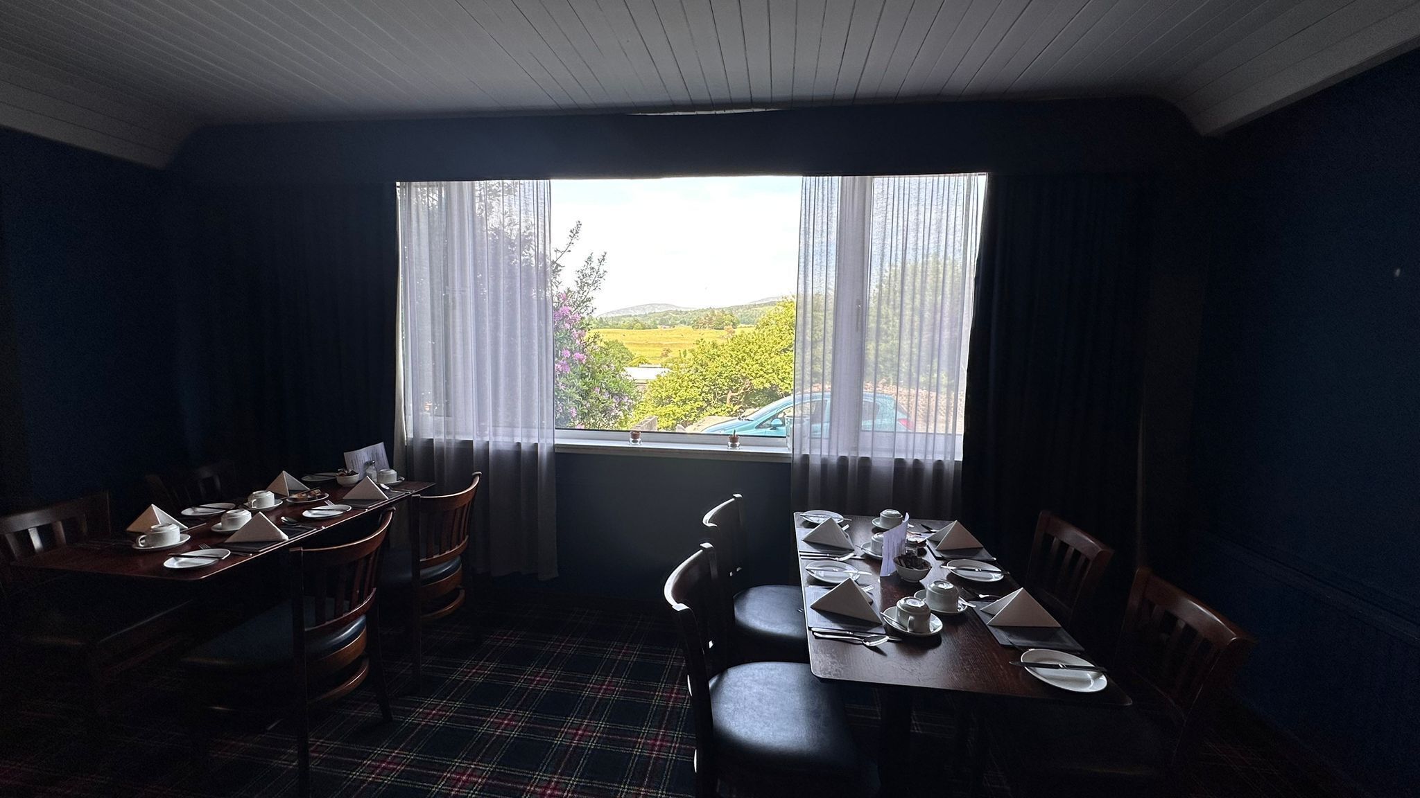 A dining room with a table set for four, a window with sheer curtains, and a plaid carpet.