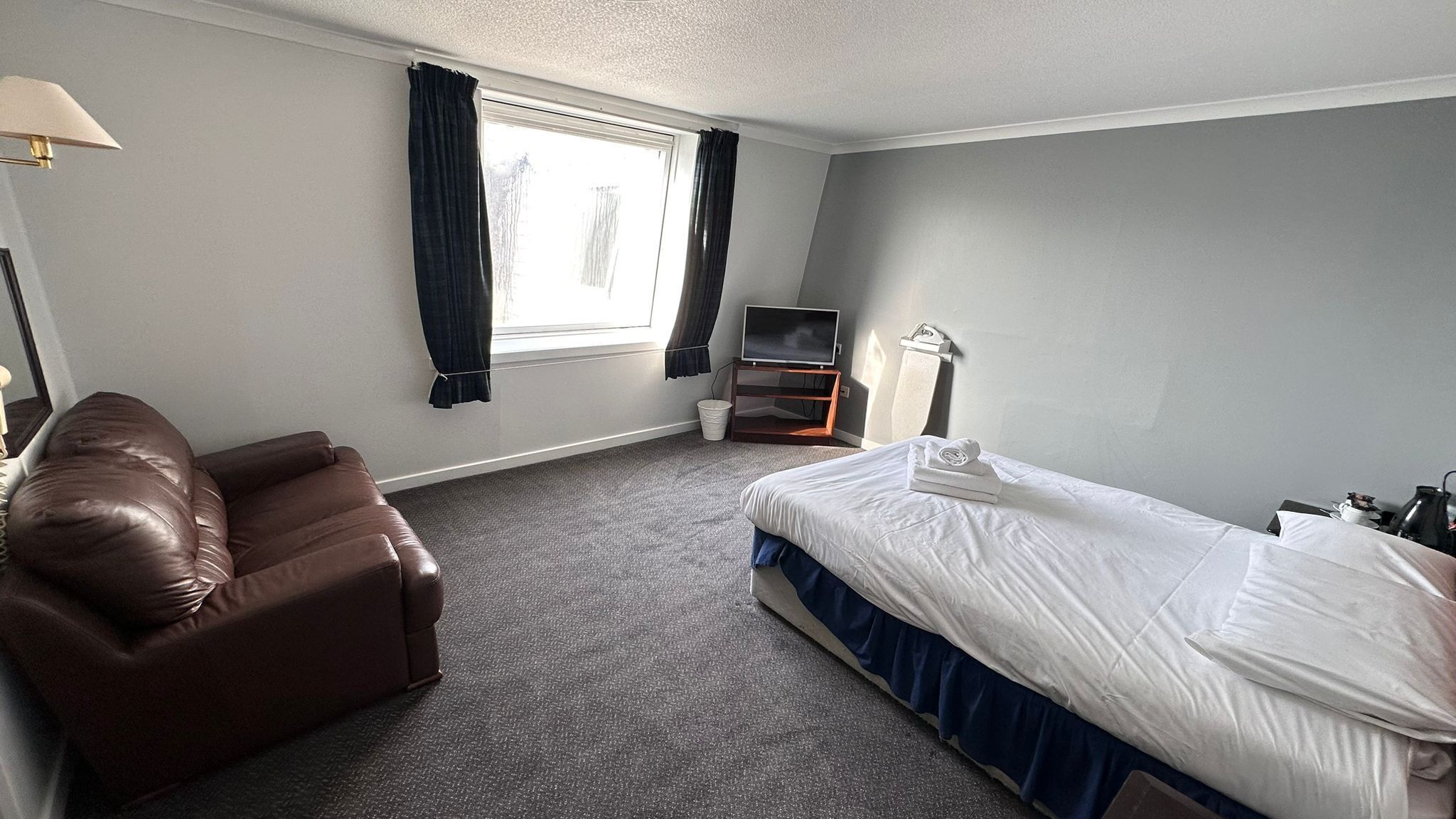 A hotel room with a single bed, a brown leather chair, a TV on a wooden stand, and a window with curtains.